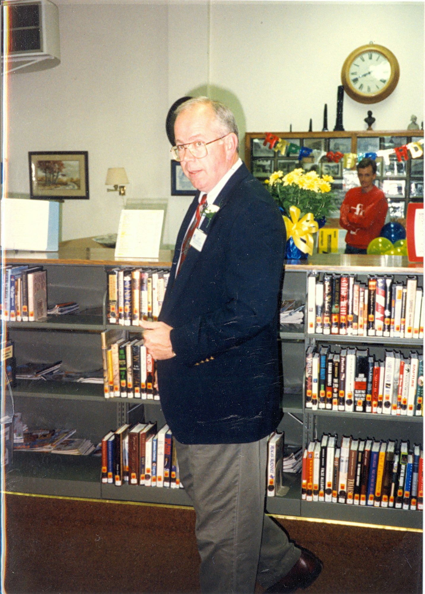 May contain: book, indoors, library, publication, shelf, adult, male, man, person, clothing, formal wear, suit, face, and head