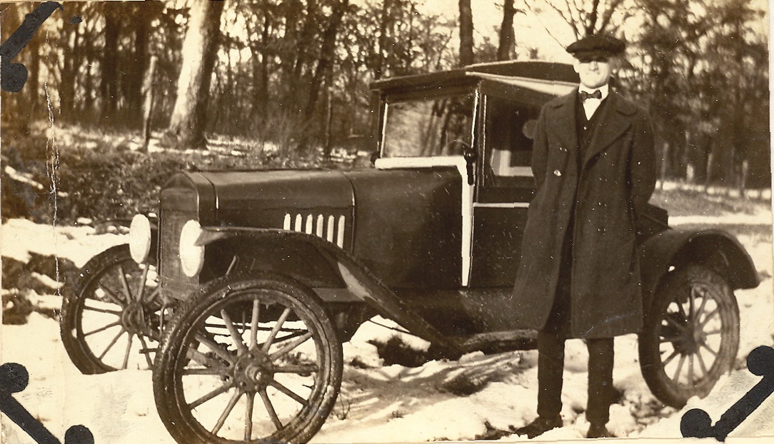 May contain: antique car, car, model t, transportation, vehicle, clothing, coat, machine, spoke, wheel, adult, male, man, and person