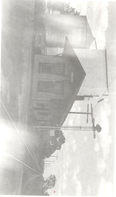 A vintage black-and-white photo of a train station, with a nearby water tower and telephone poles along the tracks.