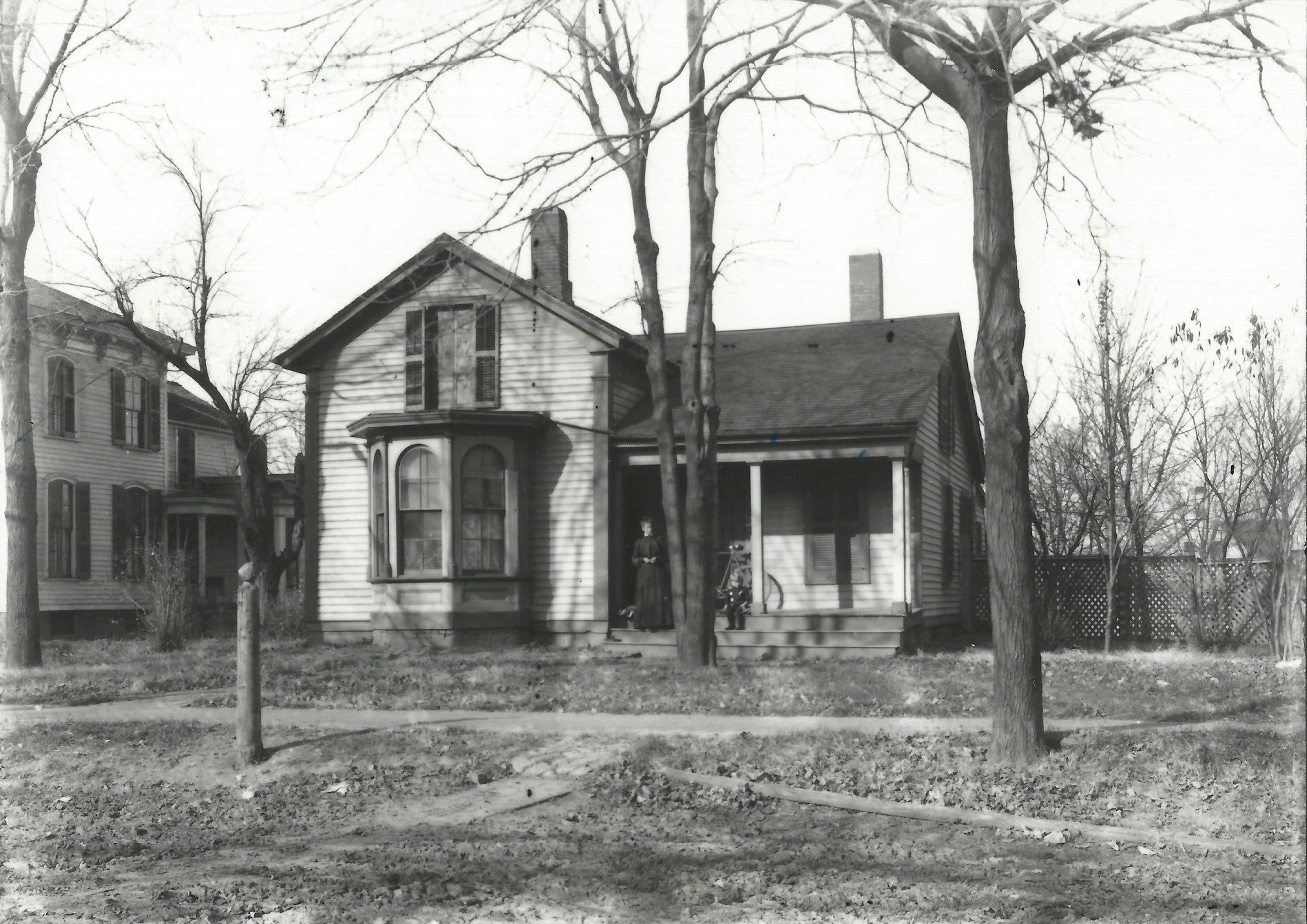 A black-and-white photo of two houses, a woman and a child on the porch, and bare trees in a residential setting.