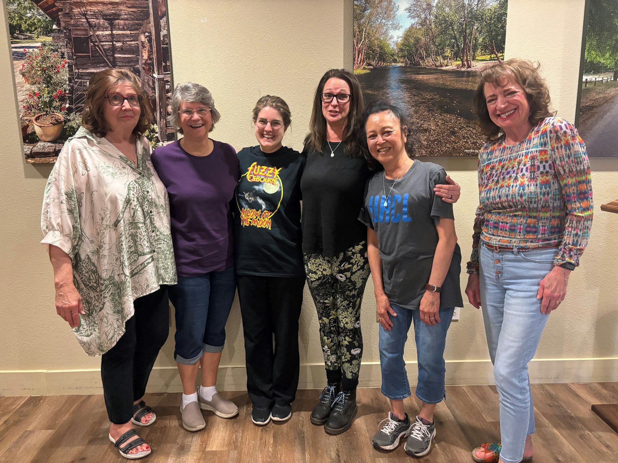 A group of six women smiling, standing side by side indoors with paintings on the wall behind them.