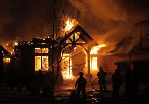 A house engulfed in flames at night with firefighters silhouetted in the foreground.