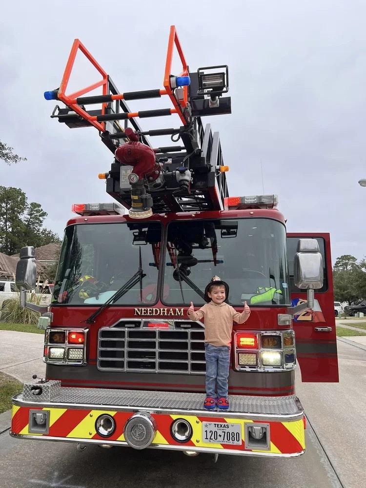 Child in front of a Needham fire truck, smiling with thumbs up, wearing a helmet.