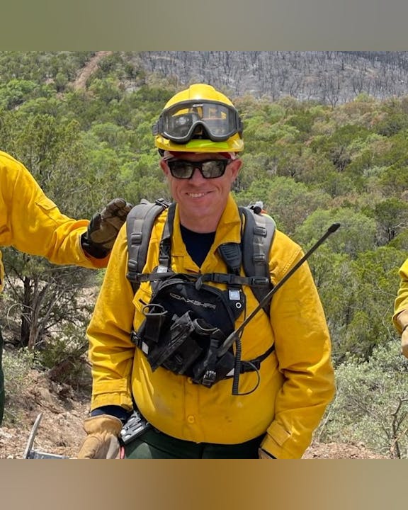 A person wearing yellow firefighting gear and sunglasses, standing outdoors in a forested area.