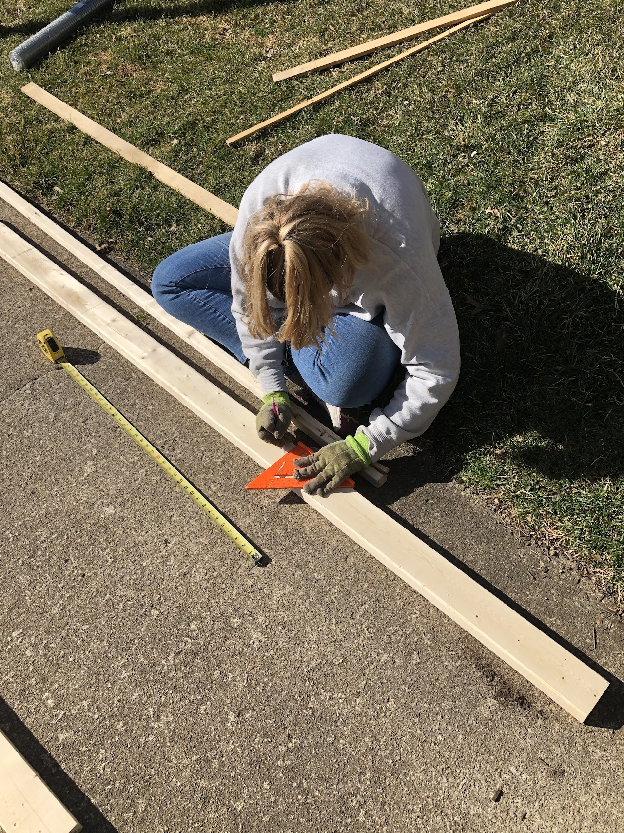 A person measuring a wooden plank with a triangular ruler and tape measure on a paved surface.