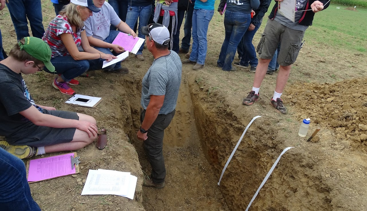 People examining a soil pit in a field, holding clipboards and notes.