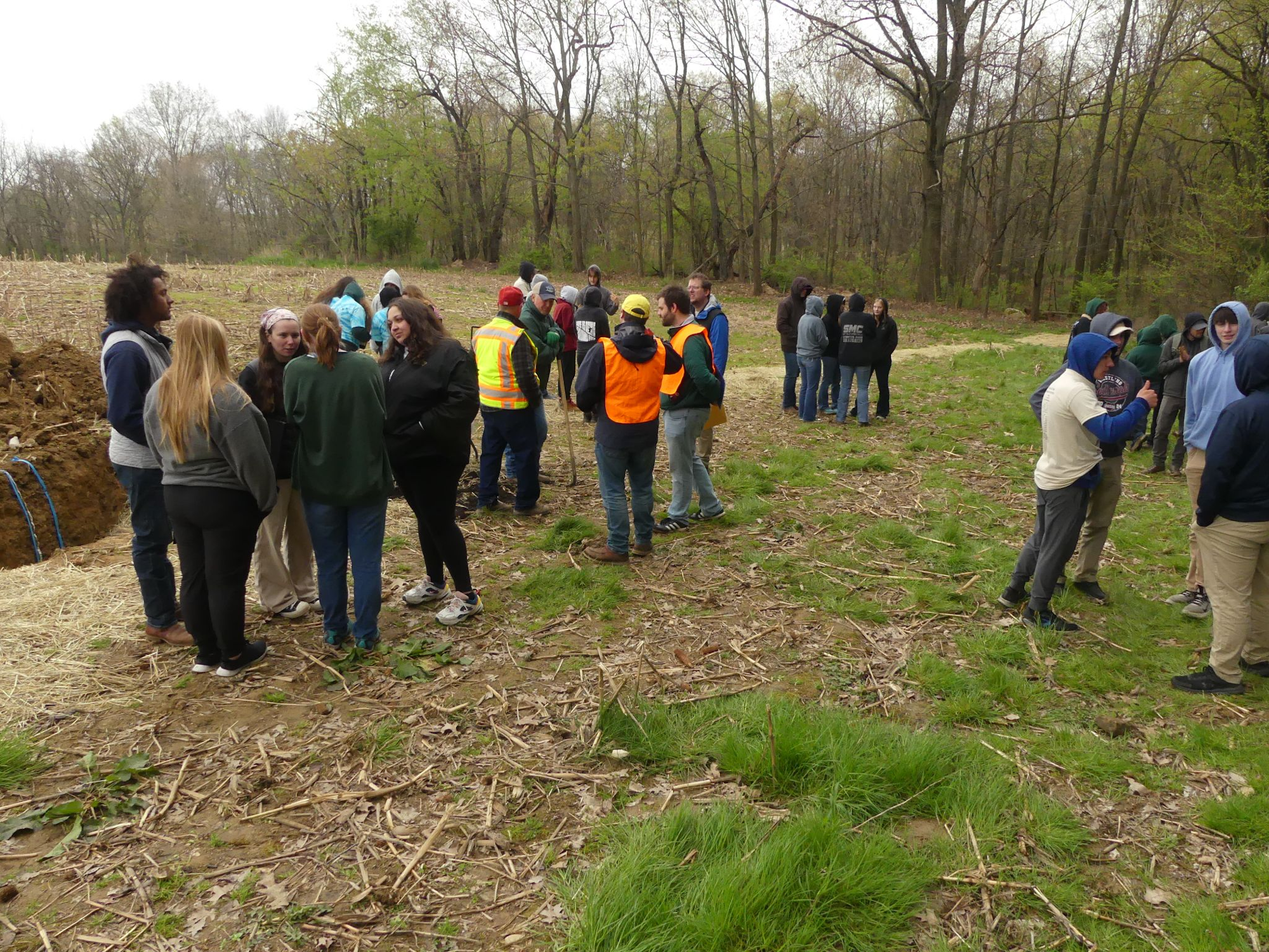 A group of people gathered outdoors, some in work vests, engaging in conversations on a grassy and wooded area.