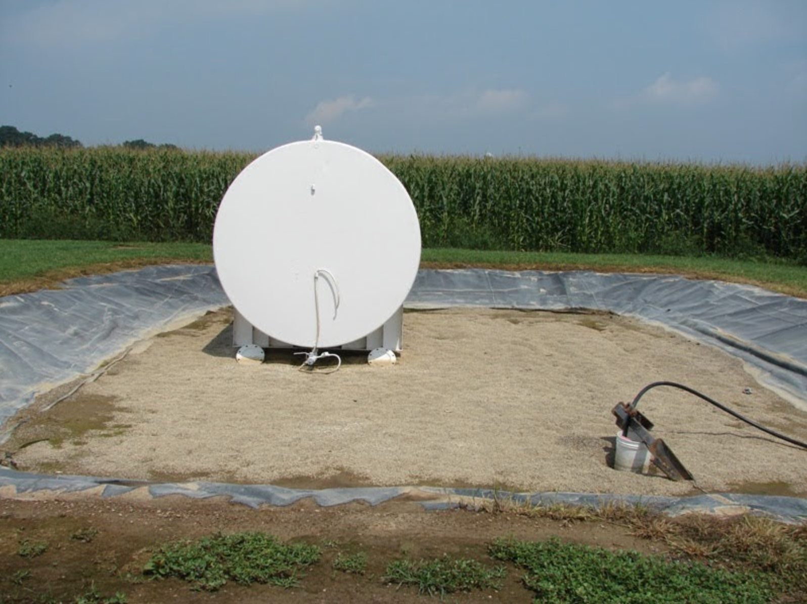 A white cylindrical tank on a prepared ground surface, with farmland and cornfield in the background.