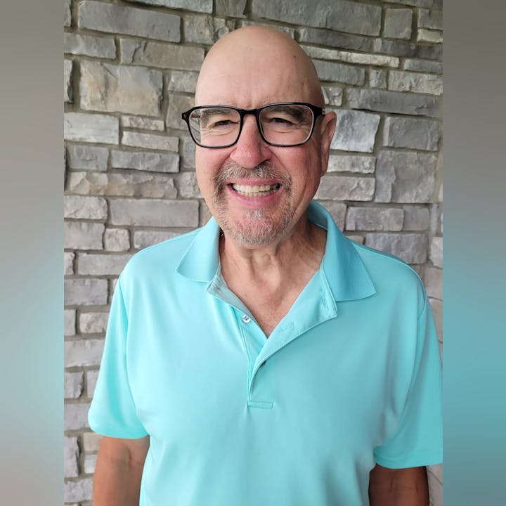 A man wearing glasses and a light blue polo shirt smiles in front of a stone wall.