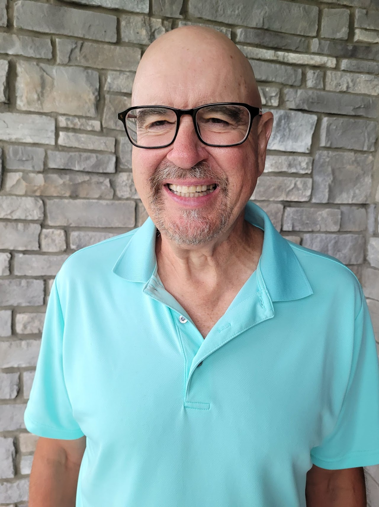 A man wearing glasses and a light blue polo shirt smiles in front of a stone wall.