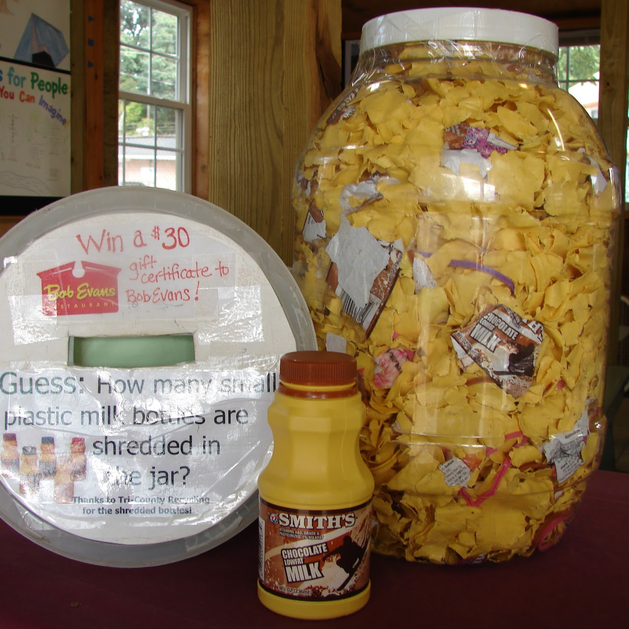 A large jar filled with shredded milk bottles, a small chocolate milk bottle, and a sign for a guessing contest to win a gift certificate.