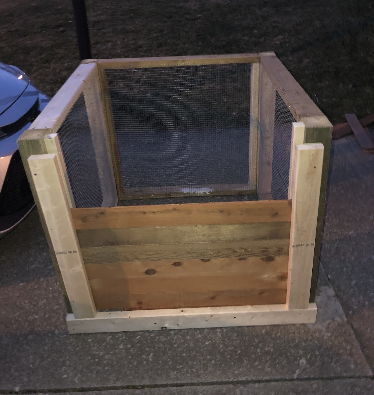A wooden and wire mesh compost bin on a concrete surface.