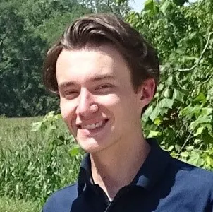 A young man in a navy polo shirt smiles and gives a thumbs-up in a green outdoor setting with trees and fields.