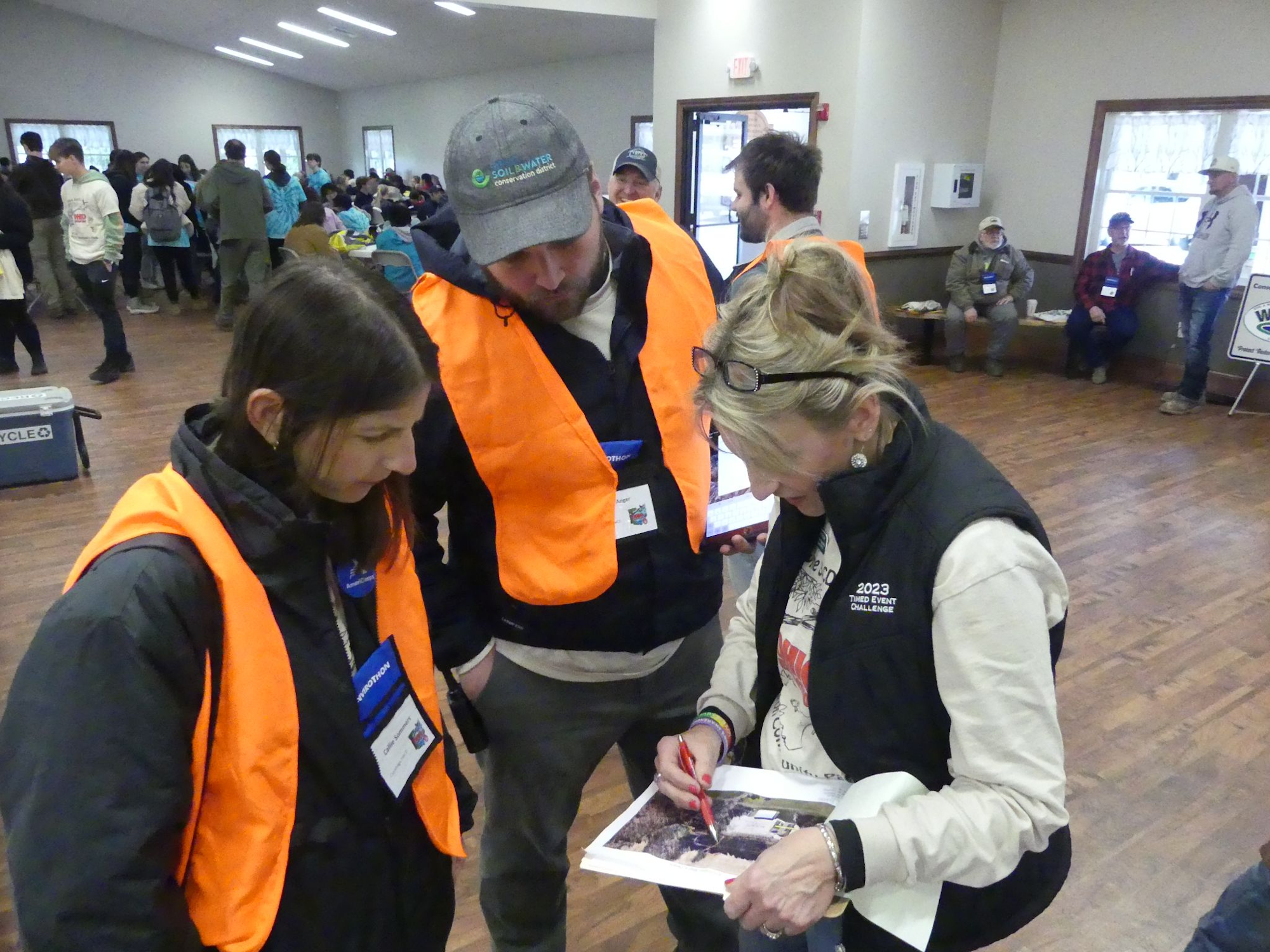 People in orange vests gathered indoors, looking at a document; others are seated and mingling in the background.