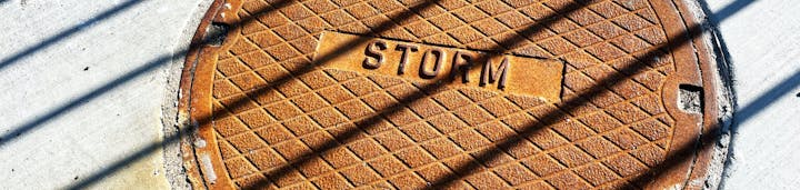 A rusty storm drain cover on concrete with shadow lines across it.