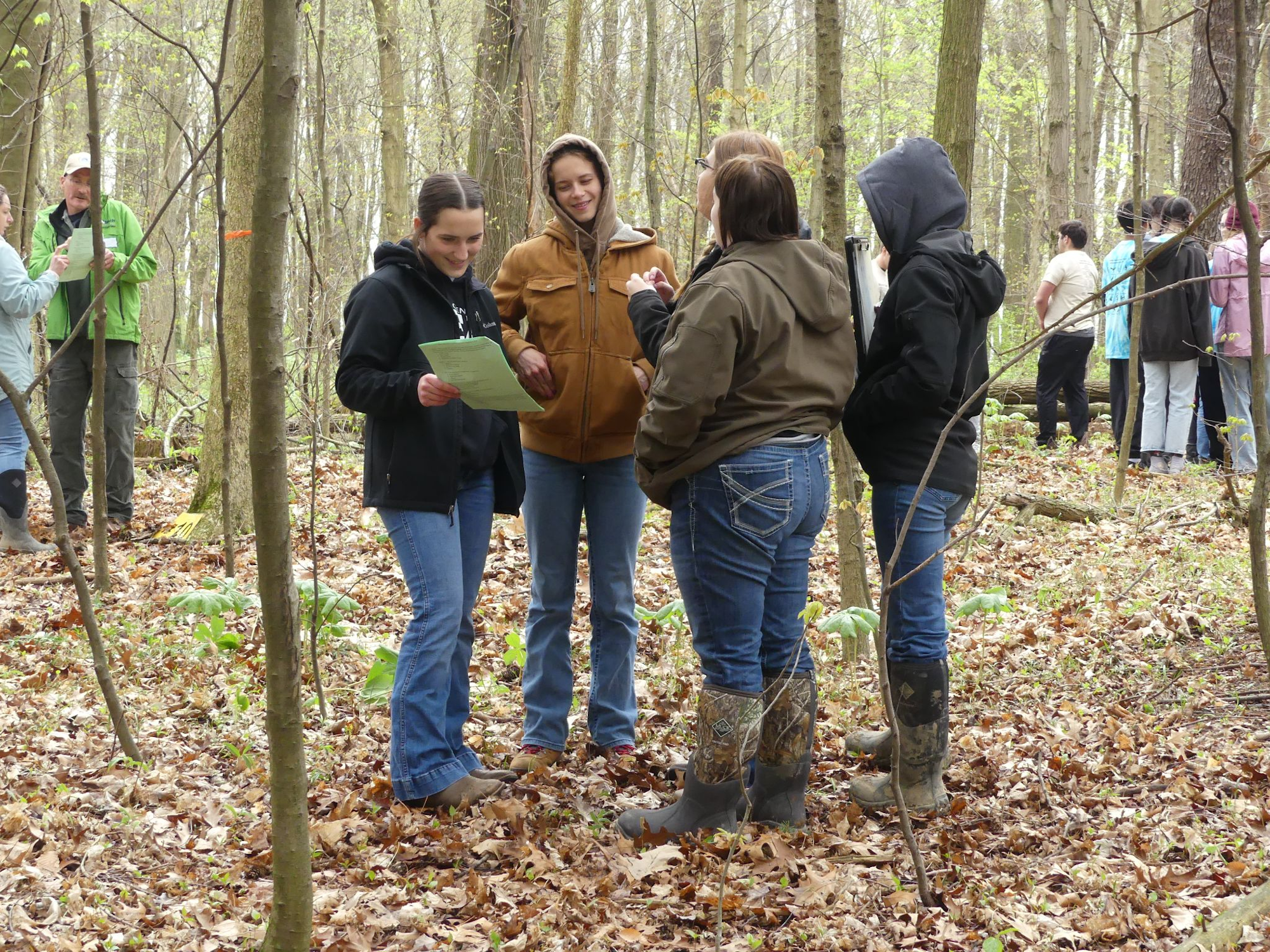 A group of people in a forest, some reading papers and wearing casual outdoor clothing and boots.