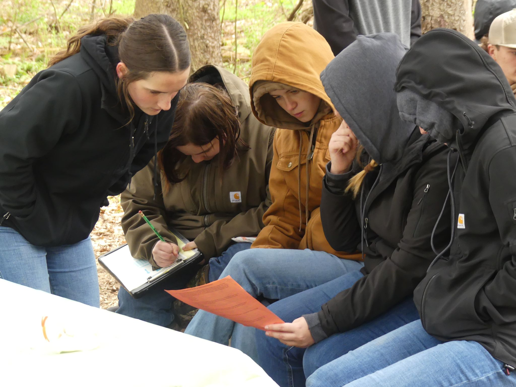 A group of people outdoors, some wearing hoodies, focus intently on papers and notes in cold weather.