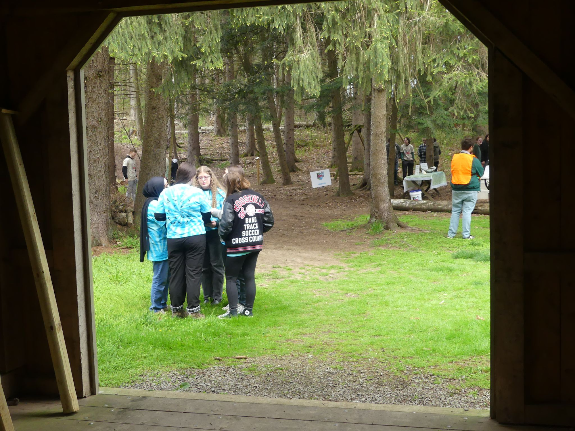 People in a wooded area, some gathered around tables; others in groups, one wearing an orange vest.