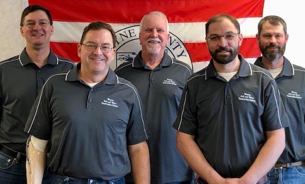 Five men wearing matching gray shirts stand in front of a flag featuring writing, likely indicating a group or organization.