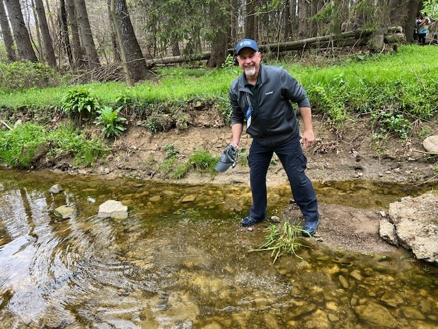 A man smiling, standing with a shoe in hand, on the edge of a shallow creek in a forested area.