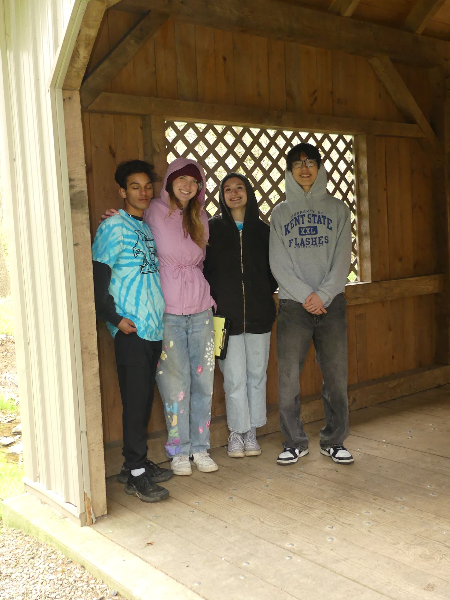 Four people wearing casual attire and hoodies stand together inside a wooden shelter.
