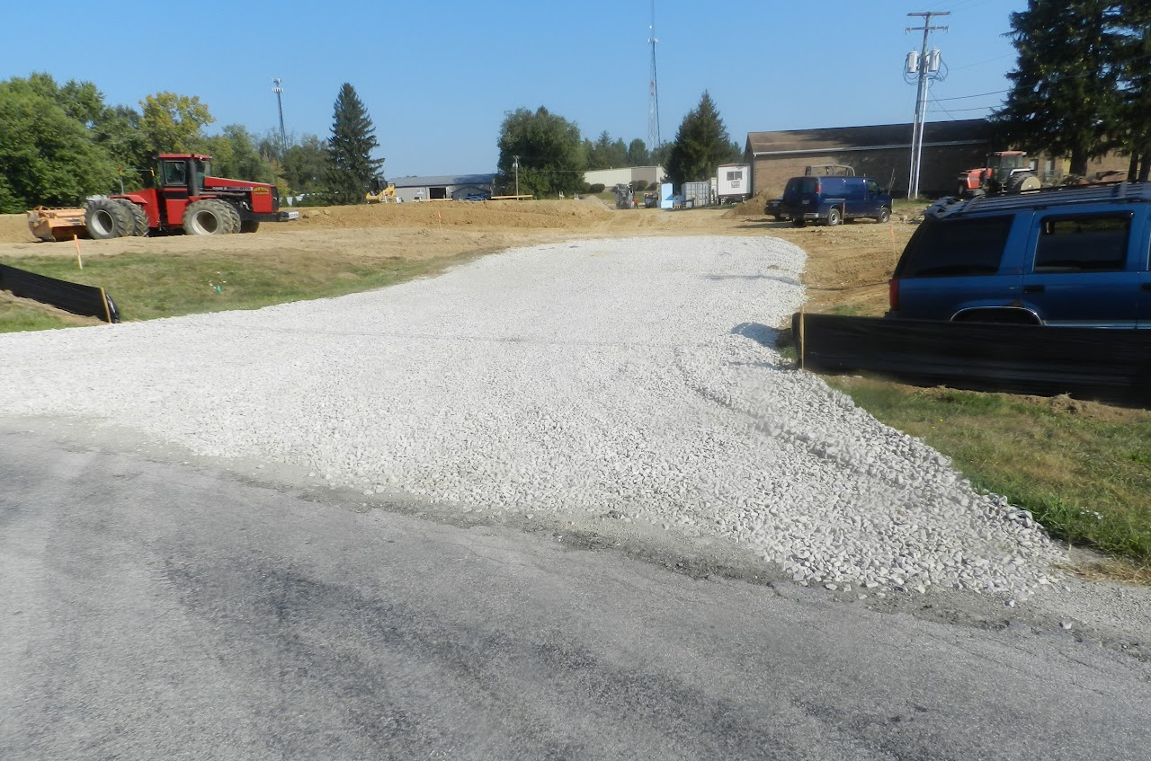 A construction site with gravel, a red tractor, vehicles, and trees in the background under a clear blue sky.