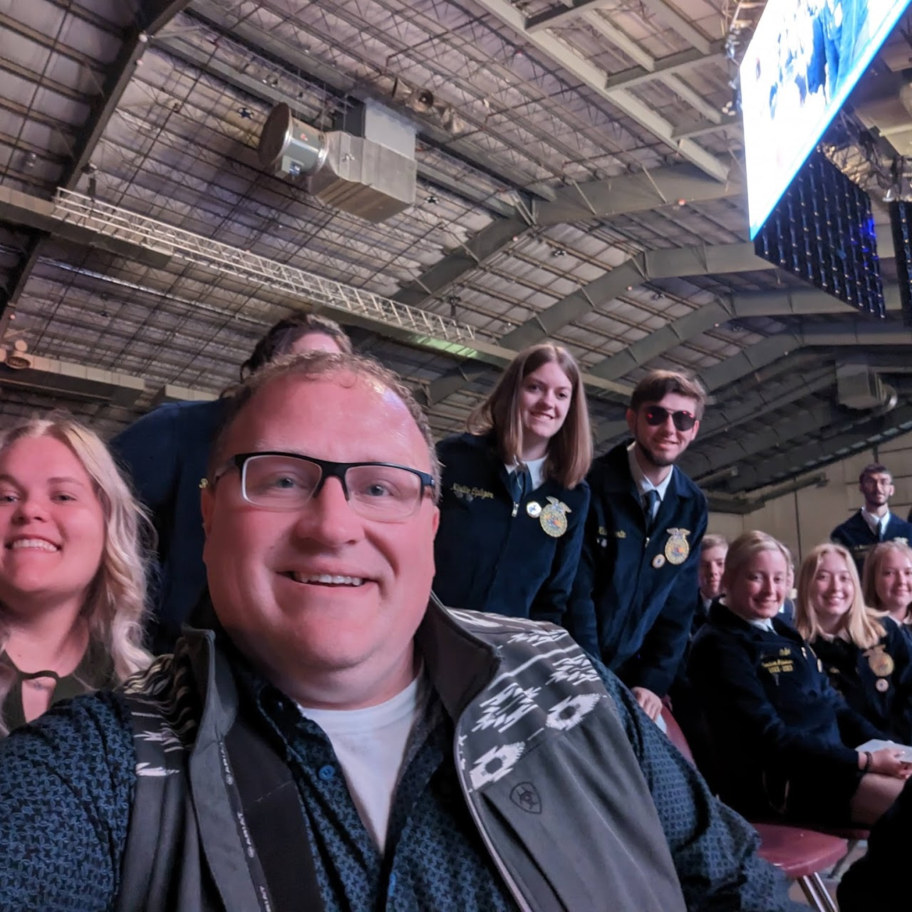 A group of smiling people posing indoors, some wearing matching jackets with patches, under a large ceiling with lights and a screen.