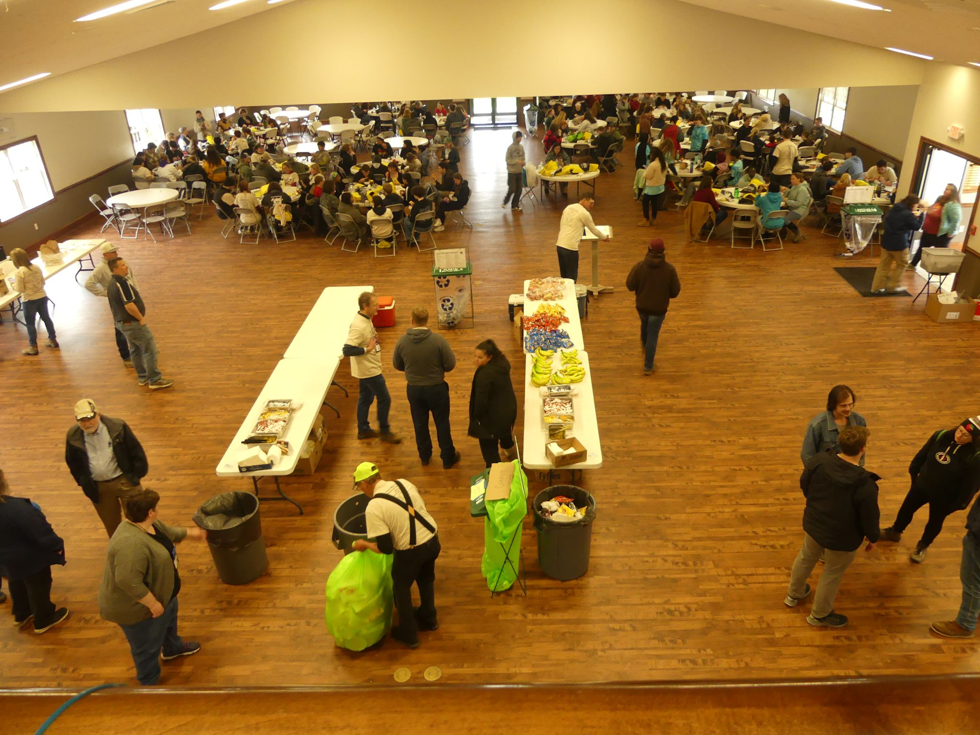 A large indoor gathering with people sitting at round tables, and a buffet table with snacks and drinks in a spacious hall.