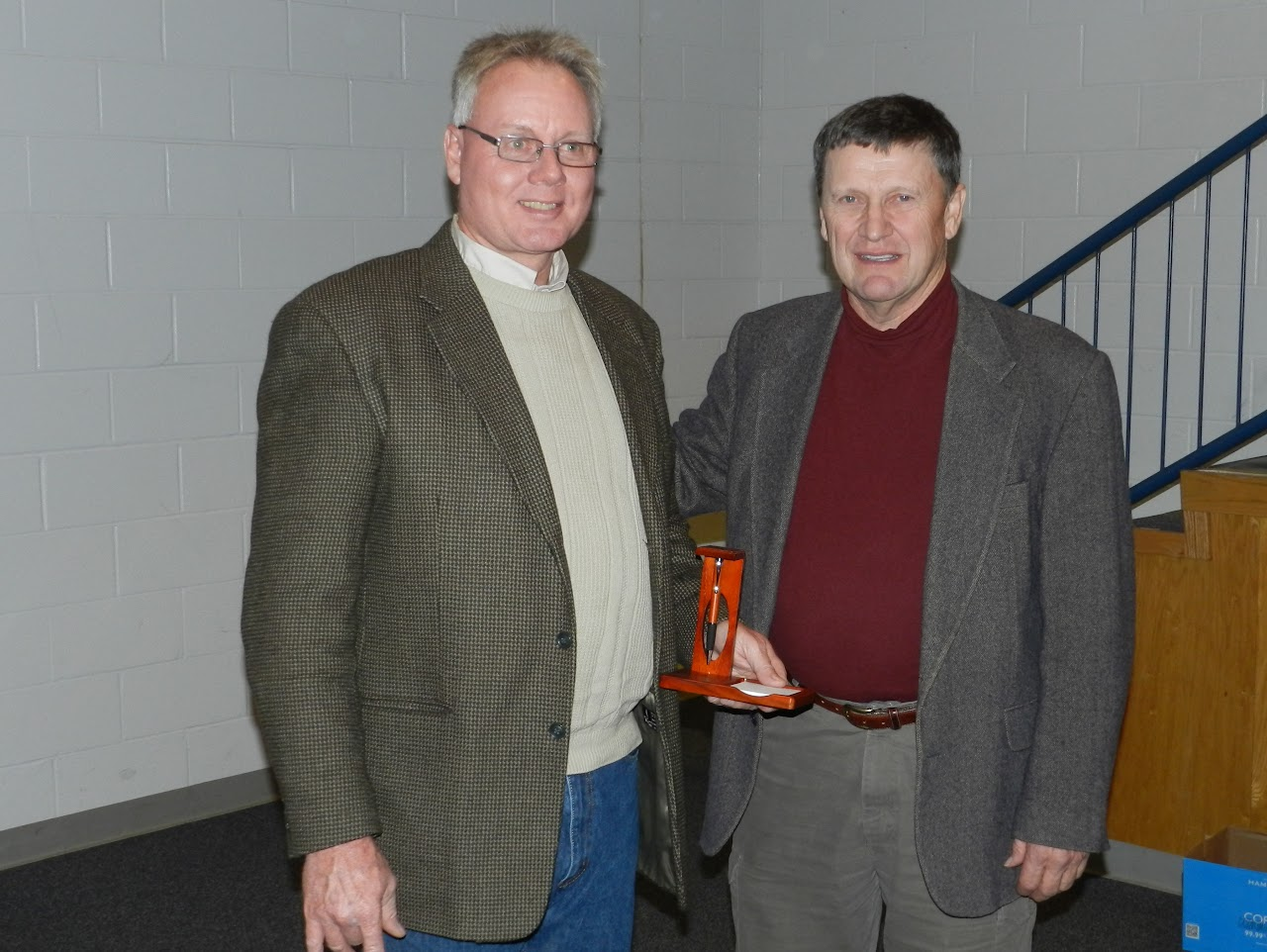 Two men in jackets standing indoors, one holding an award.