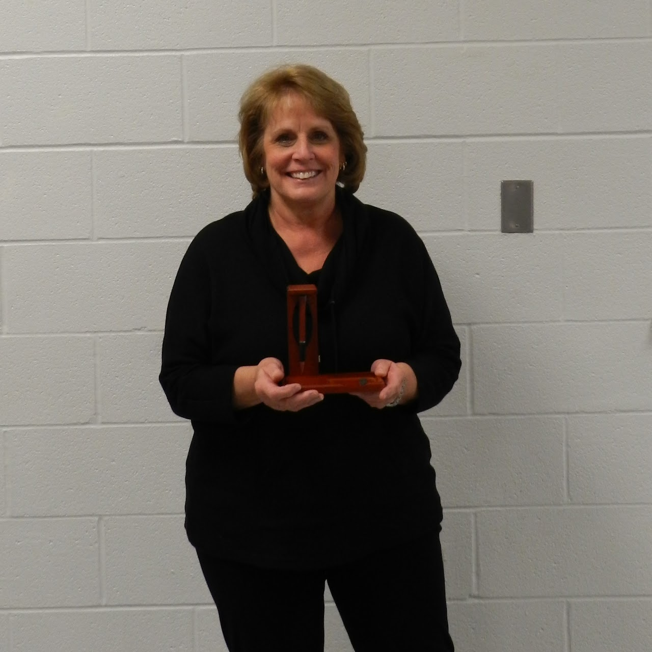 A person in a black outfit holding a small wooden object in front of a white brick wall.