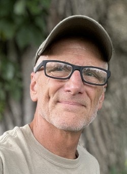 A middle-aged man with glasses and a cap, smiling softly against a natural background.