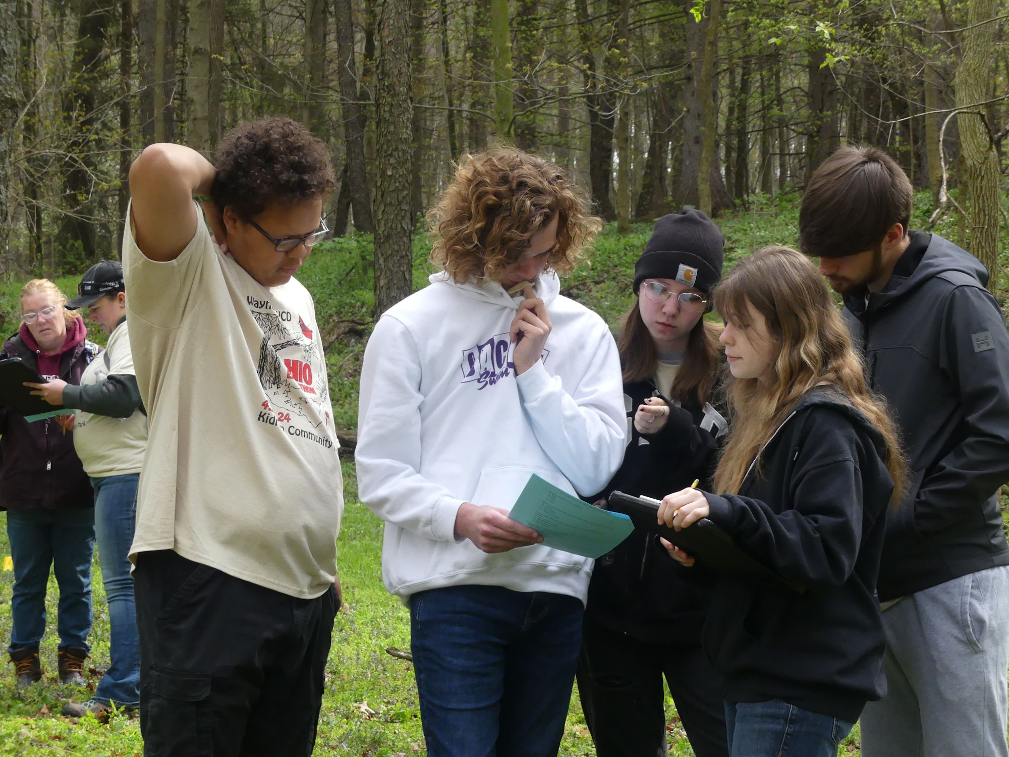 A group of people stands outdoors in a wooded area, discussing and examining papers.