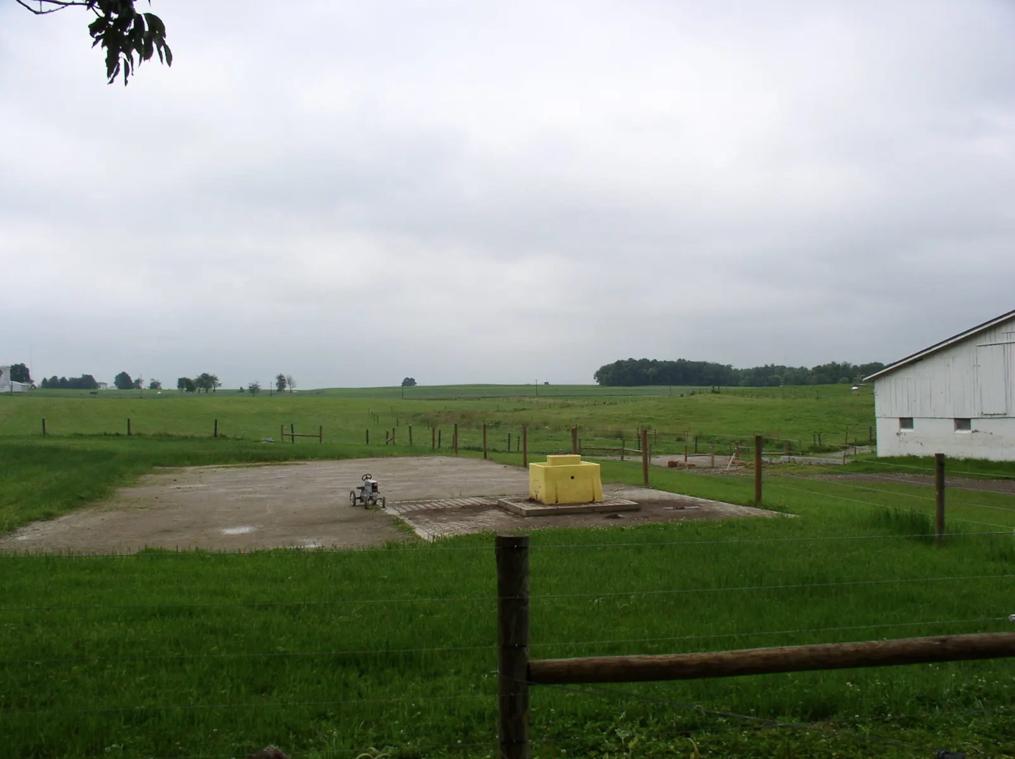 A rural landscape with green fields, a fenced area with a yellow box, and a white building under a cloudy sky.