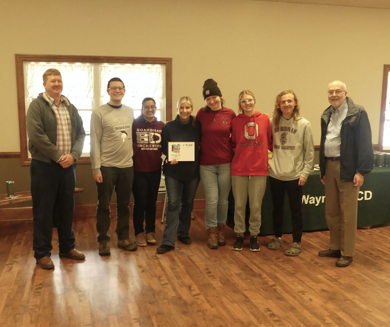 A group of people standing indoors, one person holding a "1st Place" certificate, with a banner reading "Wayne CD" in the background.