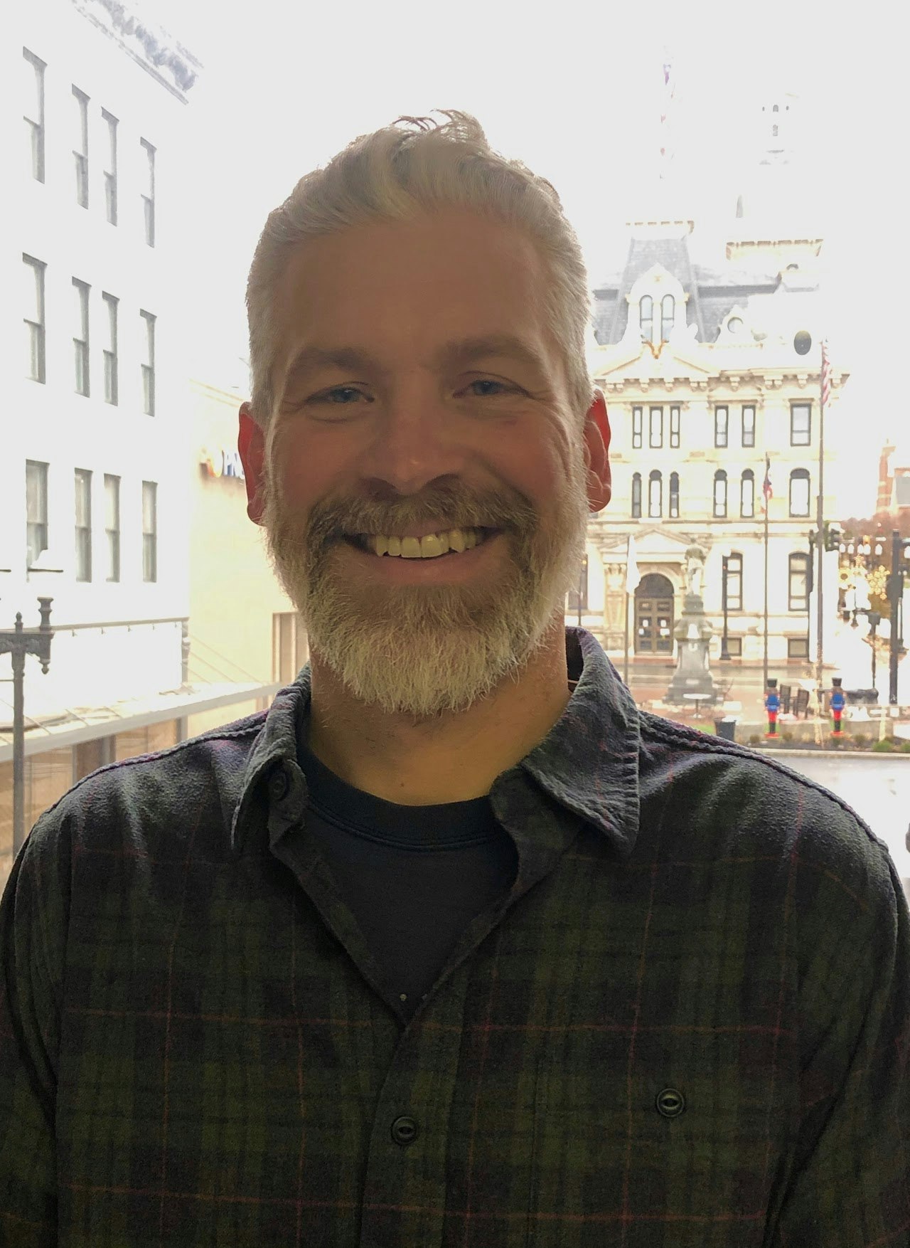A smiling man with a beard stands indoors, with a building and street scene visible outside a window behind him.