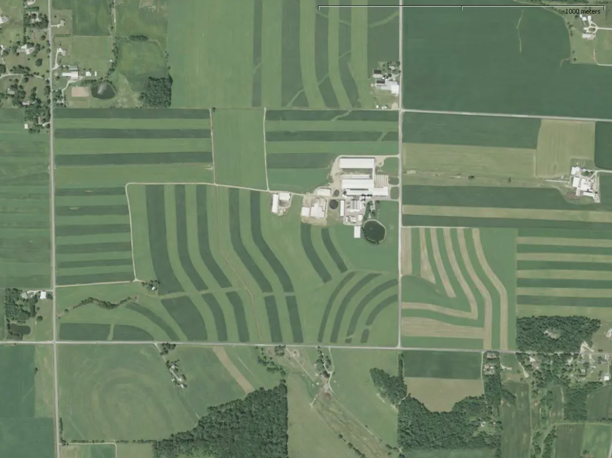 An aerial view of a landscape with farms, fields, and some buildings, featuring striped agricultural patterns.