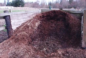 A large pile of compost or mulch in a wooden enclosure on a farm or garden area.