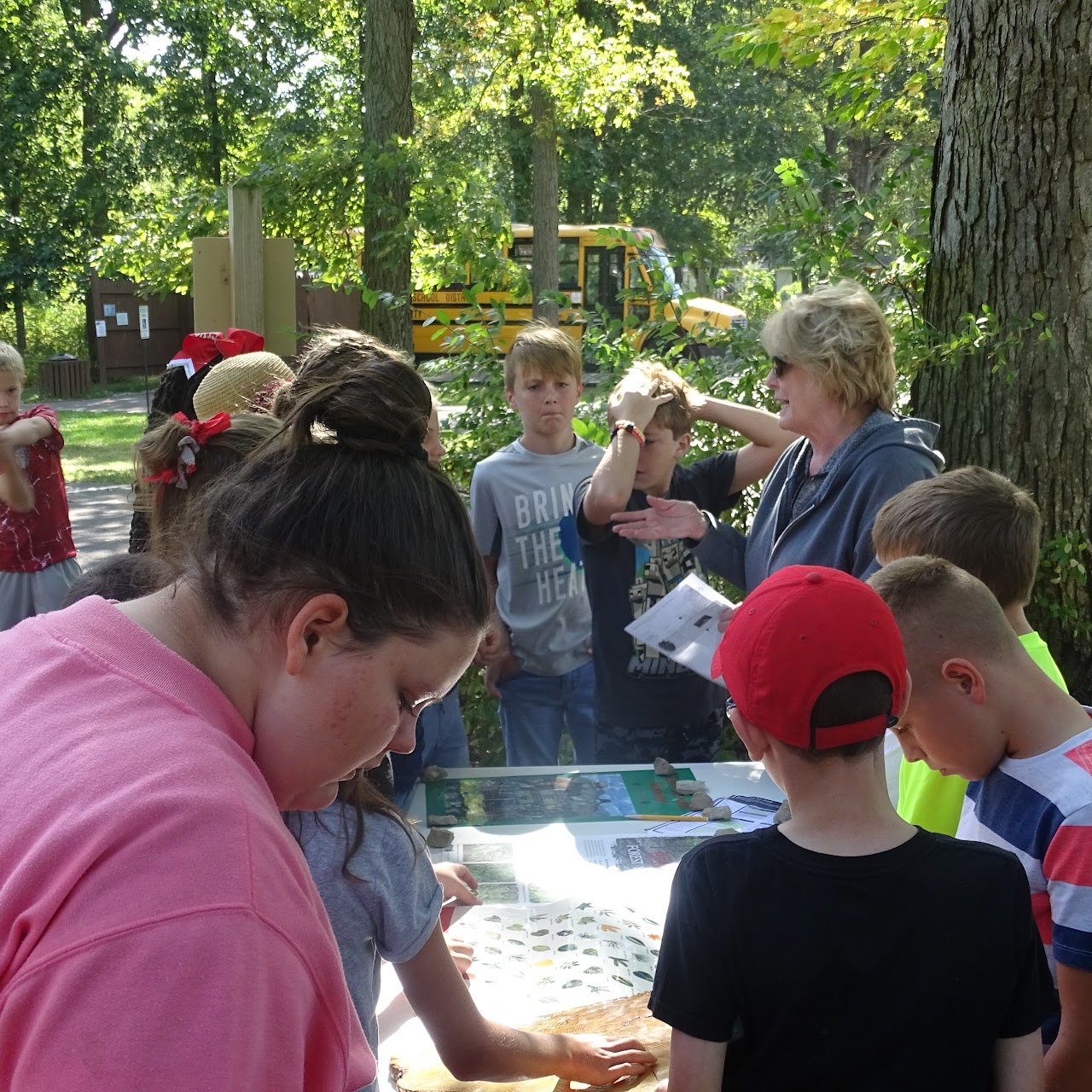 A group of kids and an adult are gathered around a table outdoors, possibly learning or examining something.