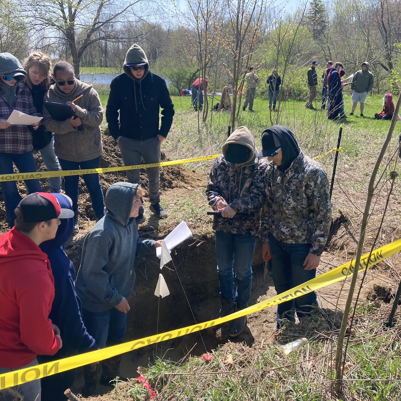 People gathered around an outdoor excavation site, examining papers. The area is marked with caution tape.