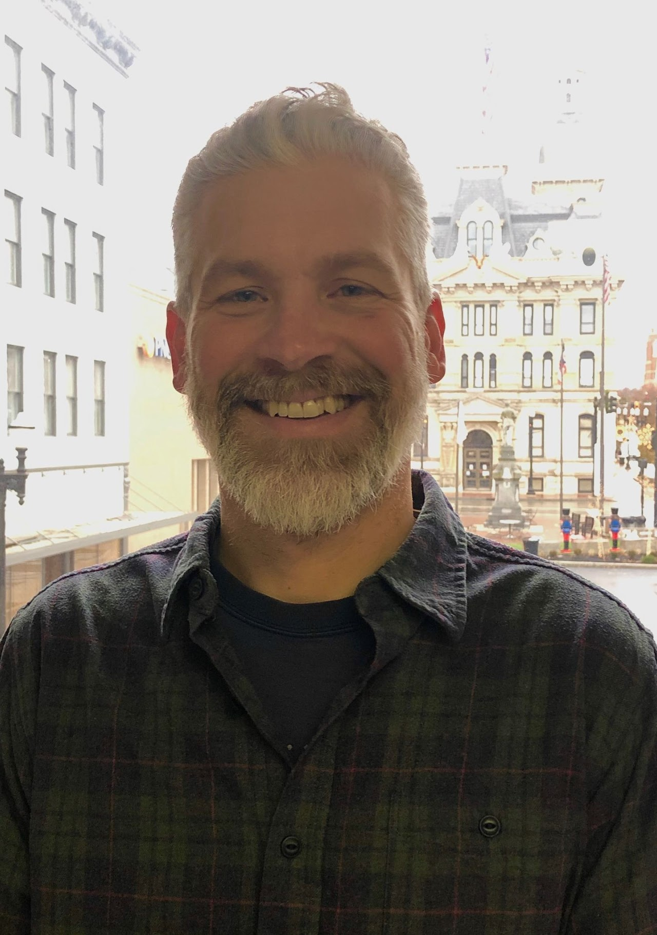 A person with a beard smiling, standing indoors with a historic building visible through the window behind them.