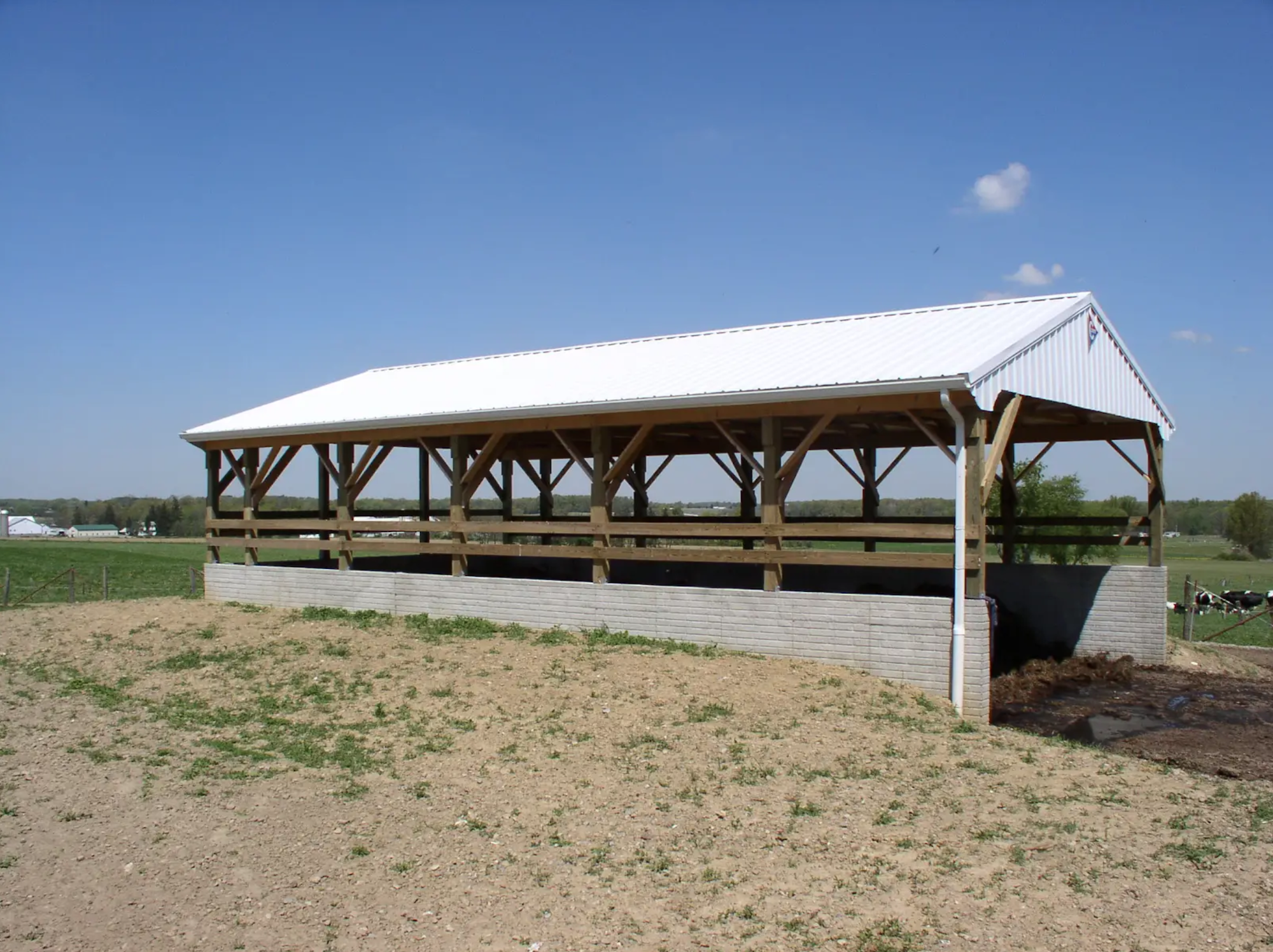 An open-sided barn with a white roof in a rural setting, surrounded by a field.