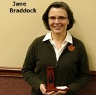 A person holding an award in a wooden case, wearing a green sweater with a leaf pin, standing indoors.