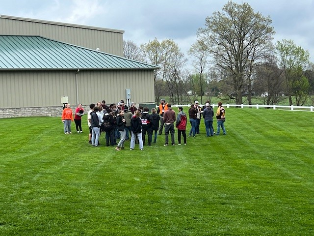 A group of people gathered on a grassy field near a building, with trees in the background.