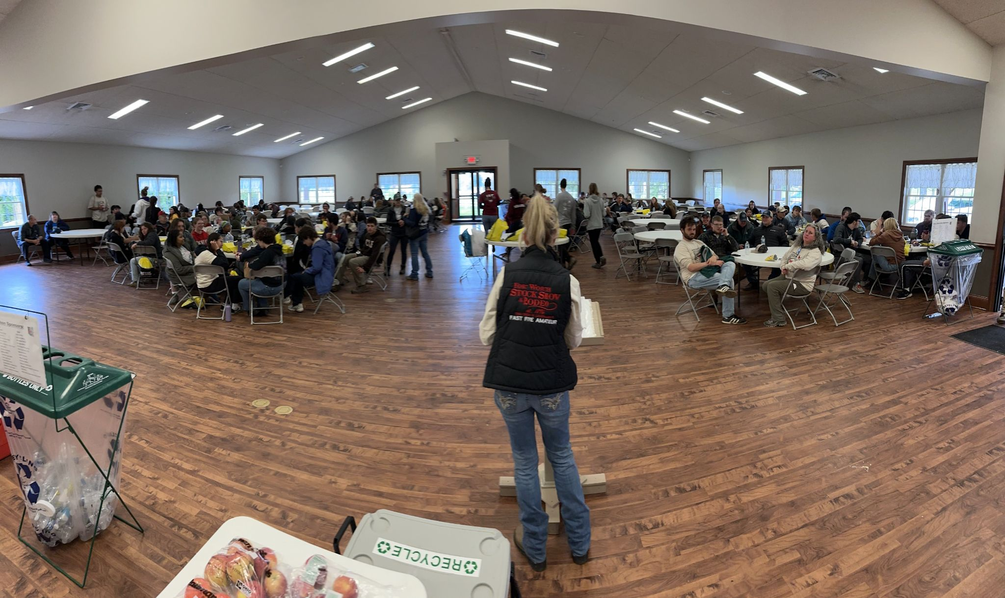 A group of people sitting at tables in a large room, with a person standing at the front, possibly giving a presentation.