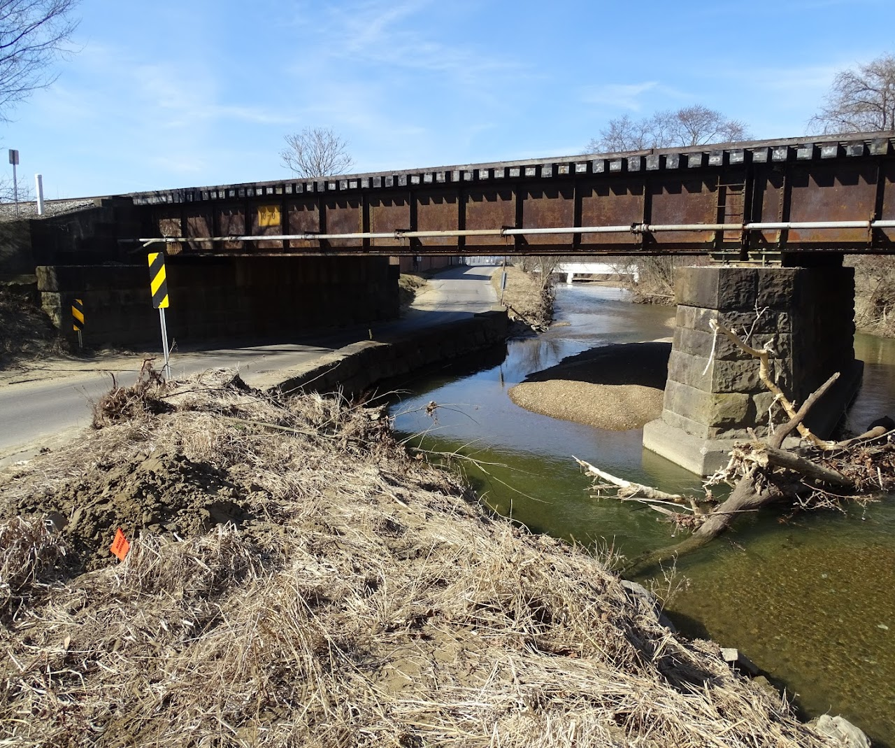 A rusty bridge over a creek, with a narrow road passing beneath it, surrounded by dry grass and bare trees.