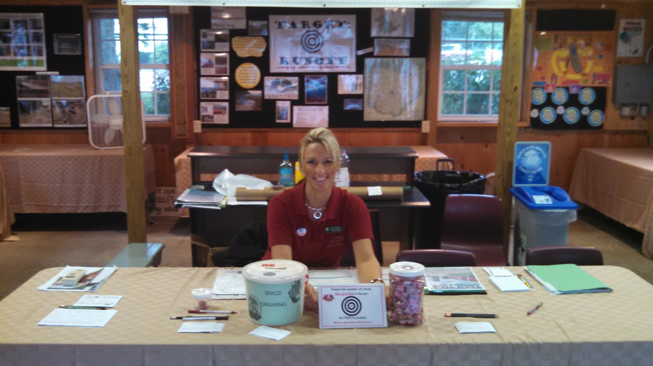 A person sits at a table in an information booth, with pamphlets, sign-up sheets, and a candy jar. Posters and exhibits are in the background.