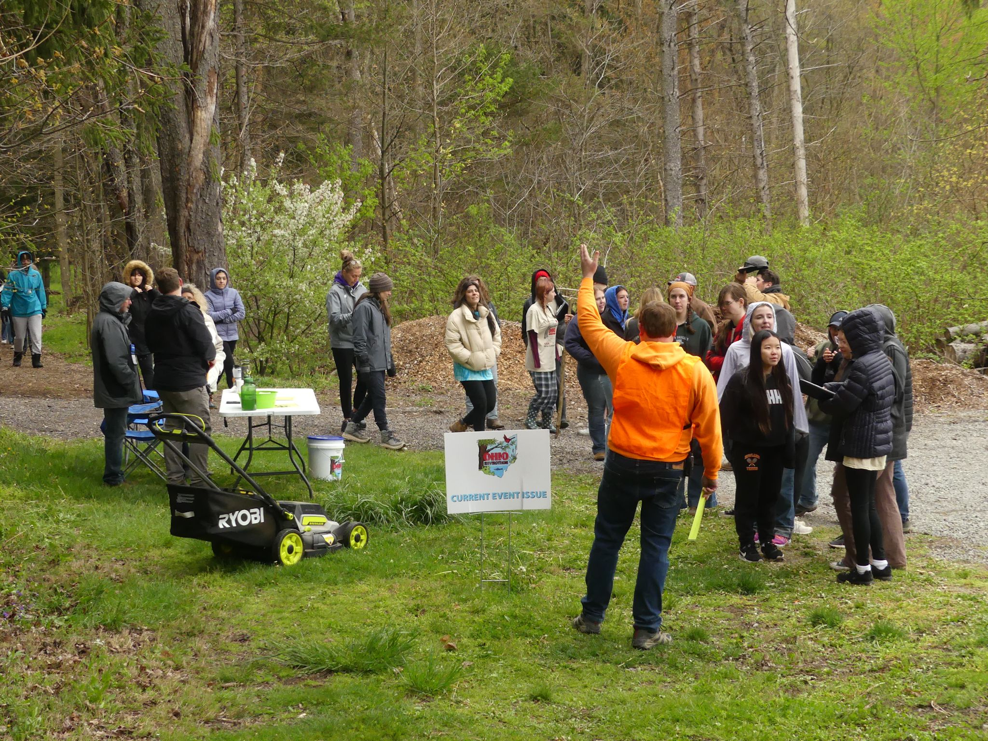 A group of people gathered outdoors, possibly for an event or tour, with a lawnmower and a sign in the foreground.