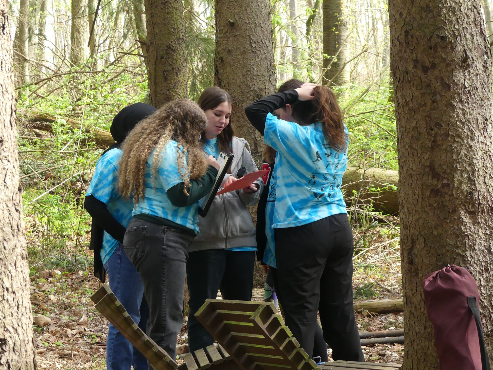 A group of people in blue tie-dye shirts is gathered in a forest, looking at a clipboard.