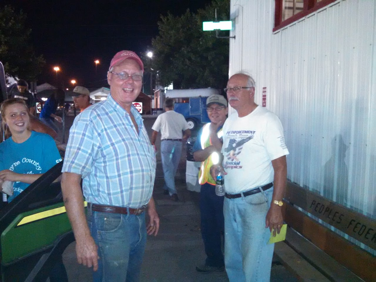 A group of people, including a man in a plaid shirt and another in a white t-shirt, smile at the camera at night near a building.