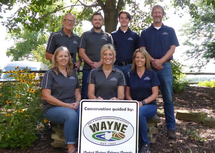 A group of people in matching shirts pose outdoors with a "Wayne" conservation sign.